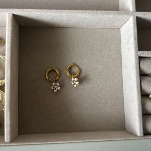 Guldiga örhängen med blommor - Söta små guldiga hoops med hängande blommor i flera färger. Blommorna har detaljer i rött, blått, grönt och lila samt en liten glittrande sten i mitten. Perfekta för dig som vill ha något färgglatt och lekfullt i öronen.