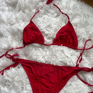 Röd bikini med knyt från NA-KD - Snygg röd bikini från NA-KD med klassisk trekantsmodell på överdelen och knyt i sidorna på underdelen. Bikinin är i mjukt syntetmaterial och har tunna band som knyts både i nacken och på höfterna. 