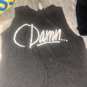 Mörkgrå ärmlös t-shirt från NLY Trend - Snygg mörkgrå ärmlös t-shirt från NLY Trend med stort vitt tryck 'Damn...' på bröstet. T-shirten har rund halsringning och är gjord i mjuk bomullsmix, perfekt för en chill och avslappnad stil.