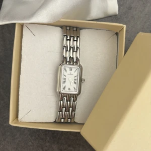 Mockberg silverfärgad rektangulär klocka - Stilren klocka från Mockberg med rektangulärt vitt ur och romerska siffror. Armbandet är i blankt silverfärgat metall med länkdesign. Inköpt för ungefär 5 månader sedan på stjärnurmakarna, säljer då jag insett att jag gillar mer guldfärgade smycken. Nypris 1899kr