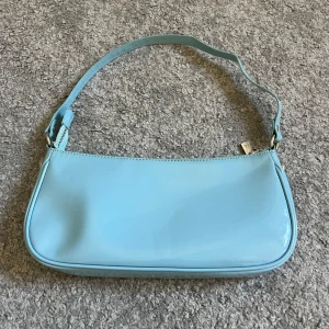 Ljusblå axelväska i glansigt material - Snygg babyblå baguetteväska från Rosemunde (Barbara Kristoffersen) i glansigt material. Väskan har en minimalistisk design med silverfärgad dragkedja och justerbart axelband. Insidan är beige med praktiska fack för småsaker. Perfekt för att lyfta din outfit med en pop av färg. Tror inte att den finns att köpa längre. Har använts men inga tecken på slitage. 