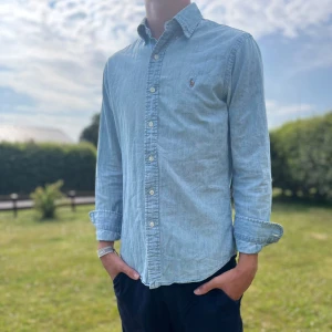 Ljusblå skjorta från Ralph Lauren - Säljer en ljusblå skjorta från Ralph Lauren i slim fit-modell. Skjortan har klassisk krage, knäppning framtill och det ikoniska broderade polospelar-loggan på bröstet. Tillverkad i mjuk bomull och har långa ärmar. Perfekt för en clean och stilren look.