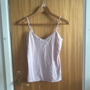 Ljusrosa linnetopp med spets från Exact - Supersöt ljusrosa linnetopp från Exact med tunna axelband och v-ringning dekorerad med spets. Perfekt för en fräsch och feminin look. Mjuk och skön bomullskänsla, passar till både jeans och kjol💗