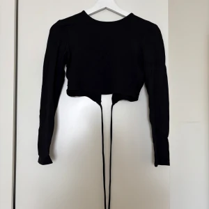Svart croppad topp från NA-KD  - Svart långärmad croppad topp från NA-KD Reborn med snörning framtill och öppen rygg. Toppens material är mjuk bomull och den har en slim passform som framhäver midjan. Perfekt för dig som gillar trendiga detaljer och stilren look.