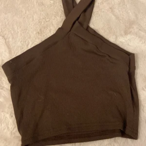 Brun ribbad one shoulder topp - Säljer en brun ribbad topp med one shoulder-design och breda axelband som korsas. Toppens material är stretchigt och sitter tight, perfekt för en trendig look. Enkel att matcha med både jeans och kjol.