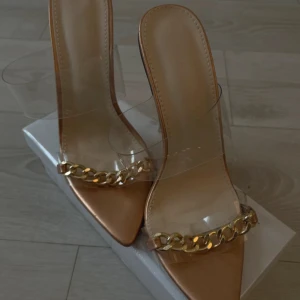 Beige klackpumps med kedjedetalj - Snygga pumps i beige med transparenta remmar och spetsig tå. Skorna har hög klack i silver och en chunky guldfärgad kedja framtill som ger en trendig look. Perfekta för dig som vill sticka ut med stilrena detaljer.