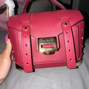 Röd handväska från Michael Kors - Snygg röd handväska från Michael Kors i skinn med guldiga metalldetaljer och nitar längs kanterna. Väskan har ett stilrent, boxigt format med handtag upptill och coolt spänne framtill. Perfekt accessoar för att lyfta din outfit.