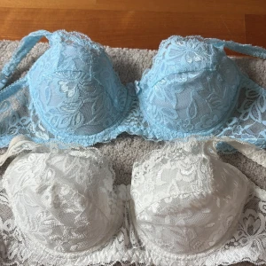 Blå och vit spetsig bh-topp - Två bh-toppar i spets, en i ljusblått och en i vitt. Båda har blommigt spetsmönster och tunna axelband. Kuporna är lätt vadderade och har dekorativ kant upptill. Perfekt för att ge en romantisk touch till outfiten. Båda för 79kr annars en för 50kr