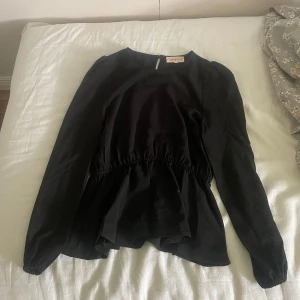 Svart blus från Only med resårmidja - Svart blus från Only med lång ärm och rund halsringning. Blusen har resår i midjan som ger en snygg siluett och lätt volang nertill. Tunt, flowy tyg som känns luftigt och skönt. Perfekt till jeans eller kjol för en stilren look.