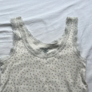 Blommig spetskantad linnetopp Brandy Melville - Supersöt vit linnetopp från Brandy Melville med små blommor i ljusrosa och grönt. Toppen har breda axelband och är dekorerad med romantisk spets runt halsringning och ärmhål. Tillverkad i mjuk bomull, perfekt för en söt och chill look.