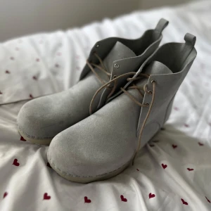 Grå mockaboots med snörning - Stilrena boots i ljusgrå mocka med klassisk snörning och rund tå. Skorna har en platt sula och diskreta sömmar längs kanten. Perfekta för dig som gillar minimalistisk och clean stil.