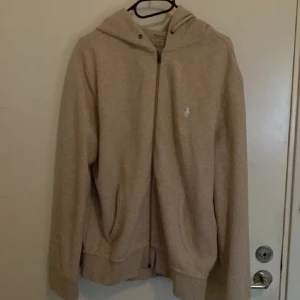 Beige hoodie från Polo Ralph Lauren - Snygg beige hoodie från Polo Ralph Lauren med dragkedja och huva. Klassisk design med två fickor framtill och diskret broderad logga på bröstet. Perfekt för chill dagar och enkel att matcha med jeans eller joggers.