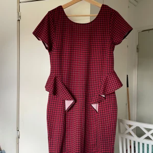 Röd och svart rutig kortklänning - Snygg kortklänning i rött och svart med rutigt mönster och coola volangdetaljer vid midjan. Klänningen har rund halsringning och korta ärmar, perfekt för dig som vill sticka ut med en unik look.