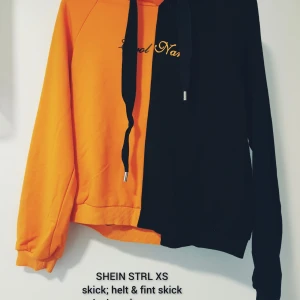 Orange och svart hoodie från SHEIN XS - Cool hoodie från SHEIN i storlek XS med halva tröjan i orange och andra halvan i svart. Framsidan har broderad text och baksidan har ett stort tryck med smiley och knivar. Tröjan har huva med snörning och långa ärmar. Perfekt statement-plagg för dig som gillar att sticka ut.  Helt skick dock små noppror