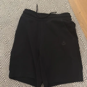 Svarta Nike tech shorts med snörning - Svarta shorts från Nike med elastisk midja och snörning. De har en stilren look, sidofickor och en bakficka med dragkedja. Perfekta för träning eller chill dagar. Materialet känns mjukt och sportigt, och Nike-loggan syns diskret framtill.
