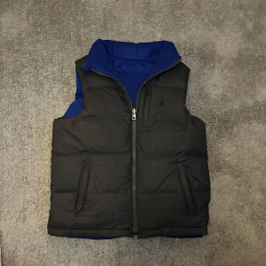Vändbar dunväst från Polo Ralph Lauren - Snygg vändbar dunväst från Polo Ralph Lauren med ena sidan svart och andra sidan blå. Västen har hög krage, två fickor på varje sida och dragkedja framtill. Perfekt för lager-på-lager och enkel att matcha med olika outfits. Nypris 1900kr,har en liten fläck
