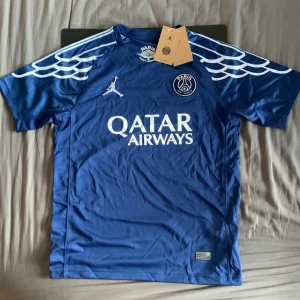 PSG x Jordan blå matchtröja 4th 24/25 - Snygg blå PSG fotbollströja från samarbetet med Jordan. Tröjan har korta ärmar, vita detaljer och vingar på axlarna. PSG-logga och Jumpman på bröstet samt Qatar Airways-tryck. Tillverkad i lätt och ventilerande funktionsmaterial.
