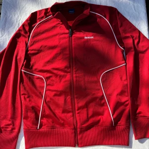 Röd vindjacka från Reebok med vita detaljer - Riktigt snygg röd vindjacka från Reebok med vita piping-detaljer längs bröstet och fickorna. Jackan har hel dragkedja framtill, ribbade muddar och Reebok-logga broderad på bröstet. Perfekt för dig som gillar sportig och clean stil.