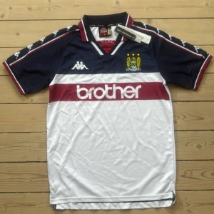 Manchester City retrotröja Kappa - Retro Manchester City fotbollströja från Kappa med klassiskt klubbmärke och Brother som sponsor. Tröjan är vit med marinblå ärmar, vinröda detaljer och Kappa-loggor längs axlarna.
