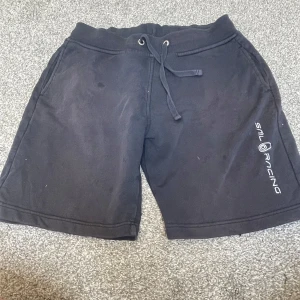 Svarta shorts från Sail Racing - Svarta shorts från Sail Racing med snörning i midjan och tryckt logga på ena benet. Ett mjukt material på insidan av bumul och med fickor både fram och bak. Perfekta för chill dagar eller sportiga aktiviteter.