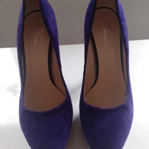 Lila mocka pumps från Rizzo, stl 36 - Snygga pumps från Rizzo i en djup lila färg, tillverkade i mjuk mocka. Skorna har hög klack och plattform för extra höjd och stil. Rund tå och klassisk siluett, perfekta för dig som vill sticka ut med färg. Made in Italy.