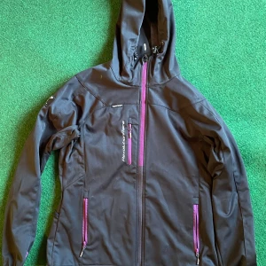 Svart hybridjacka från RVRC med huva - Svart hybridjacka från RVRC med huva och snygga lila dragkedjor. Jackan har flera praktiska fickor med dragkedja och justerbar huva med snörning. Perfekt för dig som gillar att vara ute och vill ha en stilren look. 