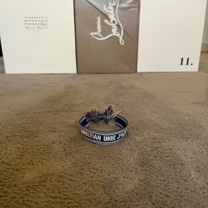 Blått armband från Christian Dior - Snyggt blått armband från Christian Dior med texten 'Christian Dior J'Adior' broderad i vitt. Armbandet har justerbar knytning med tofsar och är gjort i tyg. Perfekt accessoar för att lyfta din outfit. Kvitto finns!
