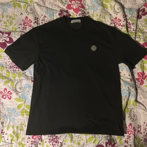 Stone Island t-shirt - Svart t-shirt från Stone Island i storlek M. Klassisk modell med rund hals och korta ärmar. På bröstet sitter en broderad Stone Island-logga i vitt och svart. Tillverkad i mjuk bomull som känns skön mot huden. Perfekt för dig som gillar stilrena och ikoniska plagg.
