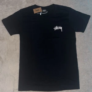 Svart Stüssy t-shirt - Svart t-shirt från Stüssy med klassisk logga i vitt på bröstet och stort färgglatt grafiskt tryck på ryggen. T-shirten har rund hals och korta ärmar, tillverkad i mjuk bomull. Perfekt för dig som gillar streetwear och vill sticka ut med cool design.