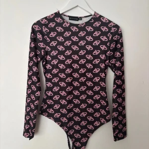 Svart body med rosa mönster från PrettyLittleThing - Cool svart body från PrettyLittleThing med lång ärm och rund hals. Toppens hela yta är täckt av ett rosa grafiskt mönster med PLT-loggor. Tight passform och stretchigt material som sitter snyggt på kroppen. Perfekt till jeans eller kjol för en trendig look.