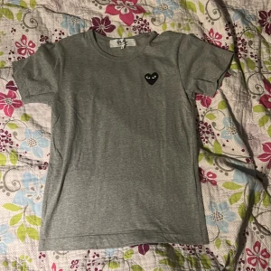 Grå t-shirt Comme des Garçons Play - Grå t-shirt från Comme des Garçons Play med det ikoniska svarta hjärtat med ögon broderat på bröstet. Klassisk rund hals och korta ärmar. Skön och stilren modell i mjuk bomull, perfekt för en clean och chill look.