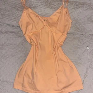 Persikofärgad linnetopp med stjärndetalj - Snygg persikofärgad linnetopp med smala, justerbara axelband och v-ringning. Toppen har en liten svart stjärndetalj framtill och är figurnära i modellen. Perfekt för dig som gillar en enkel men ändå unik look.