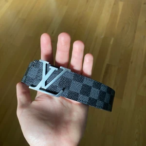rutigt svart bälte - Snyggt svart bälte  med klassiskt rutigt mönster och stor logga i silver på spännet. Bältet är gjort i skinn och har en stilren och exklusiv känsla som lyfter vilken outfit som helst.