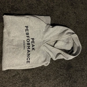 Grå hoodie från Peak Performance - Snygg ljusgrå hoodie från Peak Performance med svart logga tryckt på bröstet. Klassisk modell med huva och mjukt material, perfekt för chill dagar. Passar dig som gillar enkel och stilren streetwear.
