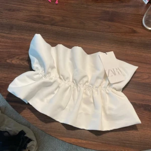 Vit bandeau tubtopp från Zara - Supersnygg vit bandeau tubtopp från Zara med volangliknande rynk längs hela toppen. Tjockare tyg med lätt glansig finish och elastisk passform. Perfekt till sommarens outfits och lätt att styla med jeans eller kjol. Liten smink fläck där bak som går bort i tvätten 