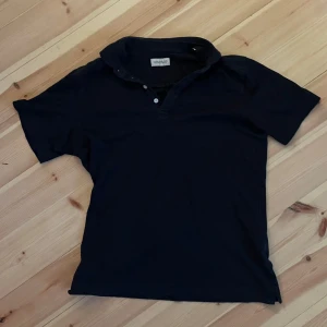 Mörkblå pikétröja från These Glory Days - Snygg mörkblå pikétröja från These Glory Days med klassisk krage och två knappar framtill. Tröjan är kortärmad och gjord i mjuk bomull, perfekt för en chill och stilren look. Passar dig som gillar enkel och tidlös stil.