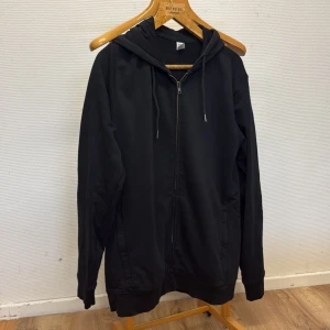 Svart hoodie med dragkedja från Clique - Svart hoodie från Clique i ekologisk bomull. Klassisk modell med huva, snörning och hel dragkedja framtill. Perfekt för chill dagar eller när du vill ha en enkel och stilren look. Stora fickor på sidorna och mjukt material.