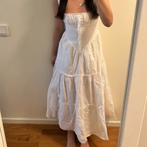 Vit långklänning med smock och volang - Superfin vit långklänning med smock upptill och volangig kjol. Klänningen är ärmlös och har en söt knytning vid bysten. Perfekt för sommarens alla tillfällen och ger en romantisk vibe.