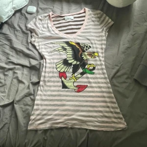 Randig Ed Hardy topp med örntryck - Säljer en randig topp från Ed Hardy i ljusrosa och grått med v-ringning. Toppen har korta ärmar och är figurnära i modellen. Perfekt för dig som gillar statement-plagg och streetstyle.