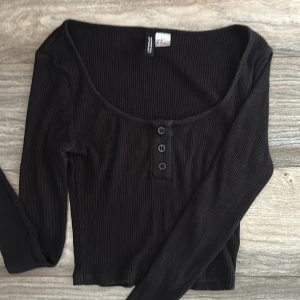 Svart ribbad topp från H&M Divided XS - Svart långärmad ribbad topp från H&M Divided i storlek XS. Toppen har rundad halsringning och tre knappar framtill som detalj. Croppad form. 