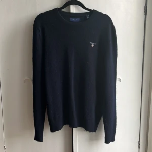 Svart stickad tröja från GANT - Klassisk svart stickad tröja från GANT med rund hals och diskret logga på bröstet. Tröjan har lång ärm och ribbade muddar vid hals, ärmslut och nederkant. Perfekt för lager-på-lager och enkel att matcha med jeans eller chinos. Är m men passar s bäst för den ör lite liten i storlek har inga defekter är lite noprig men de går att ta bort lätt säljer för ändast 300kr då nypris är 1300kr om ni har några frågor tveka inte fråga på 