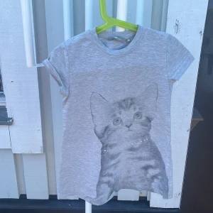 Grå t-shirt med kattmotiv från Lindex - Supersöt grå t-shirt från Lindex med ett stort tryck av en katt på framsidan. T-shirten har korta ärmar och rund hals. Materialet är mjuk bomull, perfekt för en chill och lekfull stil.