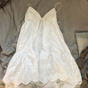 Vit somrig klänning med broderade detaljer - Superfin vit kortklänning med tunna axelband och v-ringning. Klänningen har broderade blommor och hålmönster, volangkant nertill och snörning i ryggen. Perfekt för varma dagar och ger en romantisk vibe.