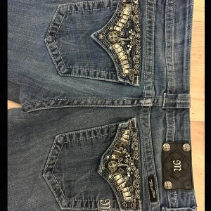 Miss Me bootcut jeans med pärldetaljer - Blå bootcut jeans från Miss Me med pärl- och metallbroderier på bakfickorna. Jeansen har låg midja, med två knappar. Backfickorna stängs inte så telefonen kan lätt läggas i bakfickan. Jeansen är märkta som storlek 29, dom passar till mig i längen men är lite små runt midjan, min storlek är M och är 174 lång. Dom har används en del men är ändå i bra skick, har bara en löss tråd på en av fickorna. 