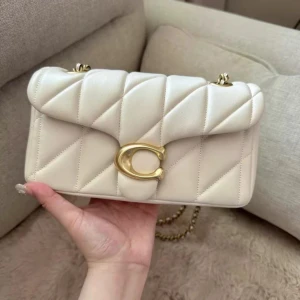 Vit quiltad axelväska från Coach - Supersnygg axelväska från Coach i ljusvit skinn med quiltad design och guldfärgade detaljer. Väskan har kedjerem, stort C-spänne framtill och flera fack inuti. Perfekt för dig som vill ha en stilren och trendig väska med lyxig känsla.