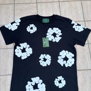 Svart blommig t-shirt från Denim Tears Medium - Svart t-shirt från Denim Tears med stora vita blommönster över hela tröjan. Klassisk rund hals och korta ärmar. Skön och stilren modell i bomull, perfekt för dig som vill sticka ut med en unik print.