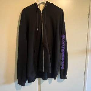 Svart hoodie med lila tryck - Svart hoodie från Junkyard i storlek XL med dragkedja, huva och snörning. Stora lila futuristiska tryck på ryggen och ärmarna med texten 'QUINTESSENCE' och grafiska detaljer. Mjuk och skön, perfekt för en streetwear-look. Köpt för ca 800kr för typ 1 år sedan men tröjan är i bra skick