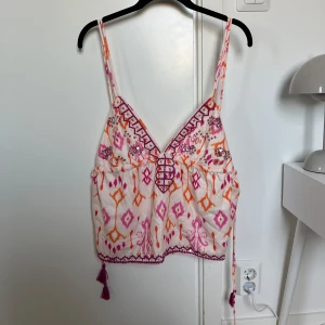 Mönstrad linnetopp med paljetter - Säljer en superfin linnetopp med smala axelband, v-ringning och snörning på sidorna. Toppen har ett färgglatt mönster i rosa, orange och lila samt små paljettdetaljer. Materialet känns lätt och luftigt, perfekt för varma dagar. Dekorativa tofsar i sidorna som gör toppen justerbar.