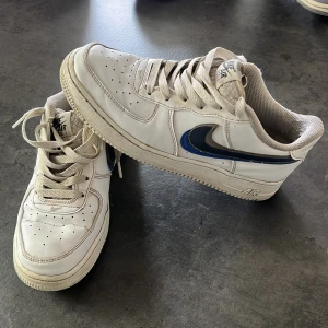 Nike Air Force 1 vita sneakers - Klassiska Nike Air Force 1 sneakers i vitt skinn med blå Swoosh på sidan. Modellen har rund tå, snörning och perforerade detaljer framtill för extra ventilation. Perfekta för dig som gillar streetstyle och tidlös design.