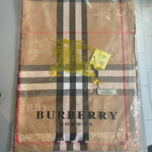 Rutig beige halsduk från Burberry - Exklusiv halsduk från Burberry i klassiskt rutigt mönster med beige, svart, vit och röd. Tillverkad i 100% kashmir och prydd med Burberrys logga och emblem. Perfekt accessoar för att lyfta din outfit med lyxig känsla.